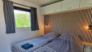 A bedroom with a bed, two bedside tables, a window with curtains, a ceiling light, and a wall-mounted shelf. - New chalet 1 on the edge of the Wadden Sea and Amstelmeer with private terrace! (Anna Paulowna)
