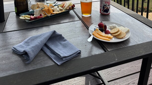 A table set outdoors with a scenic view, featuring a bottle, a glass of beer, plates of cheese and fruit, and a can of beer.