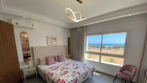 A bedroom with a bed, a chair, a nightstand, a lamp, a window with a view, and an air conditioning unit. - Haut Standing apartment in a prime location: just 5 minutes from the beach, with sea views (Mahdia)