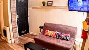 A living room with a brown sofa, a black coffee table, a TV mounted on the wall, and a door with a black handle. - Charming Jawn in Fabulous Fishtown — W/D, AC, Private Patio & Fast WiFi (Philadelphia)