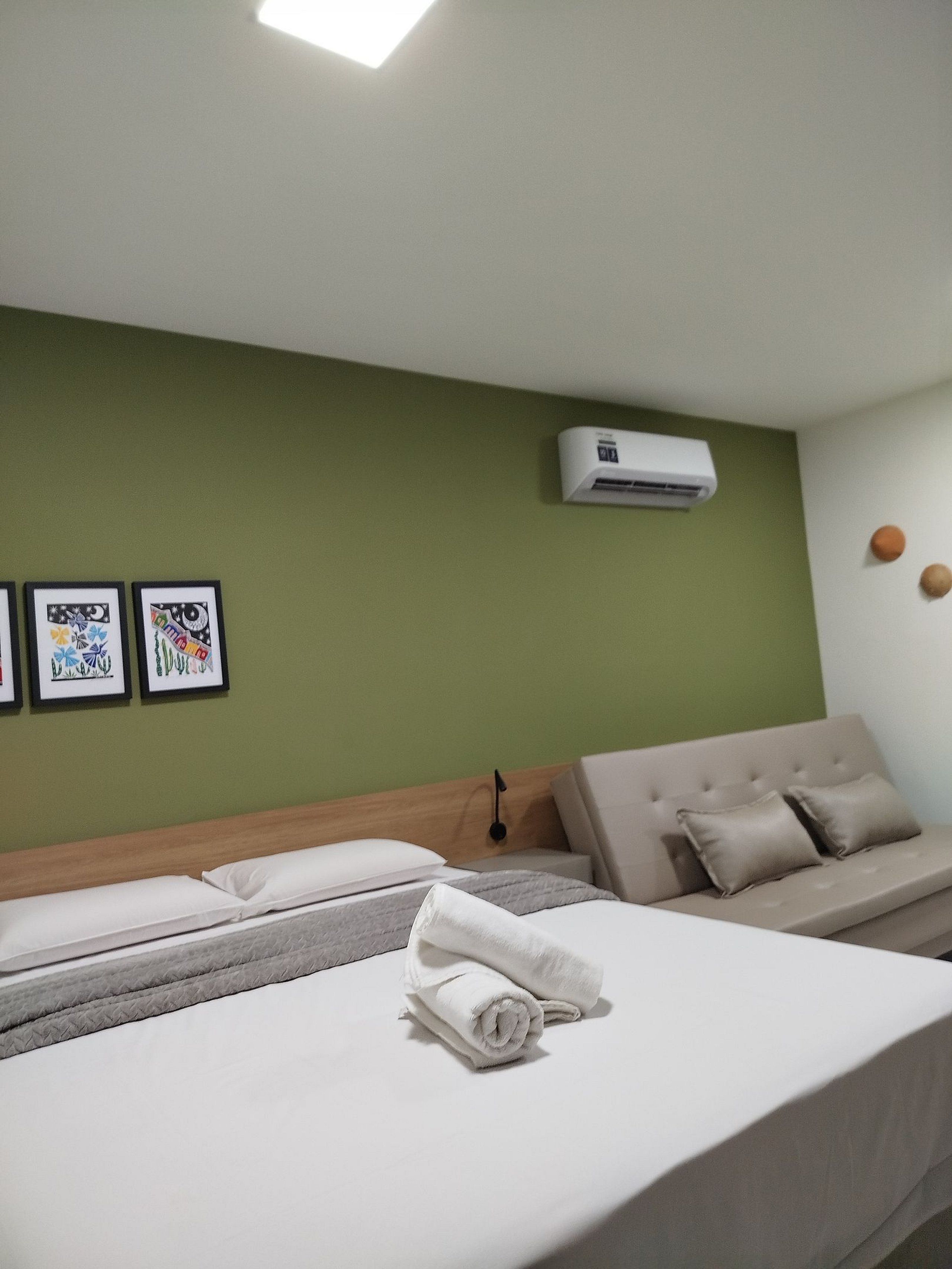 A hotel room with a bed, a sofa, an air conditioning unit, and three framed artworks on the wall.