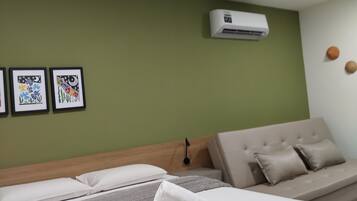 A hotel room with a bed, a sofa, an air conditioning unit, and three framed artworks on the wall.