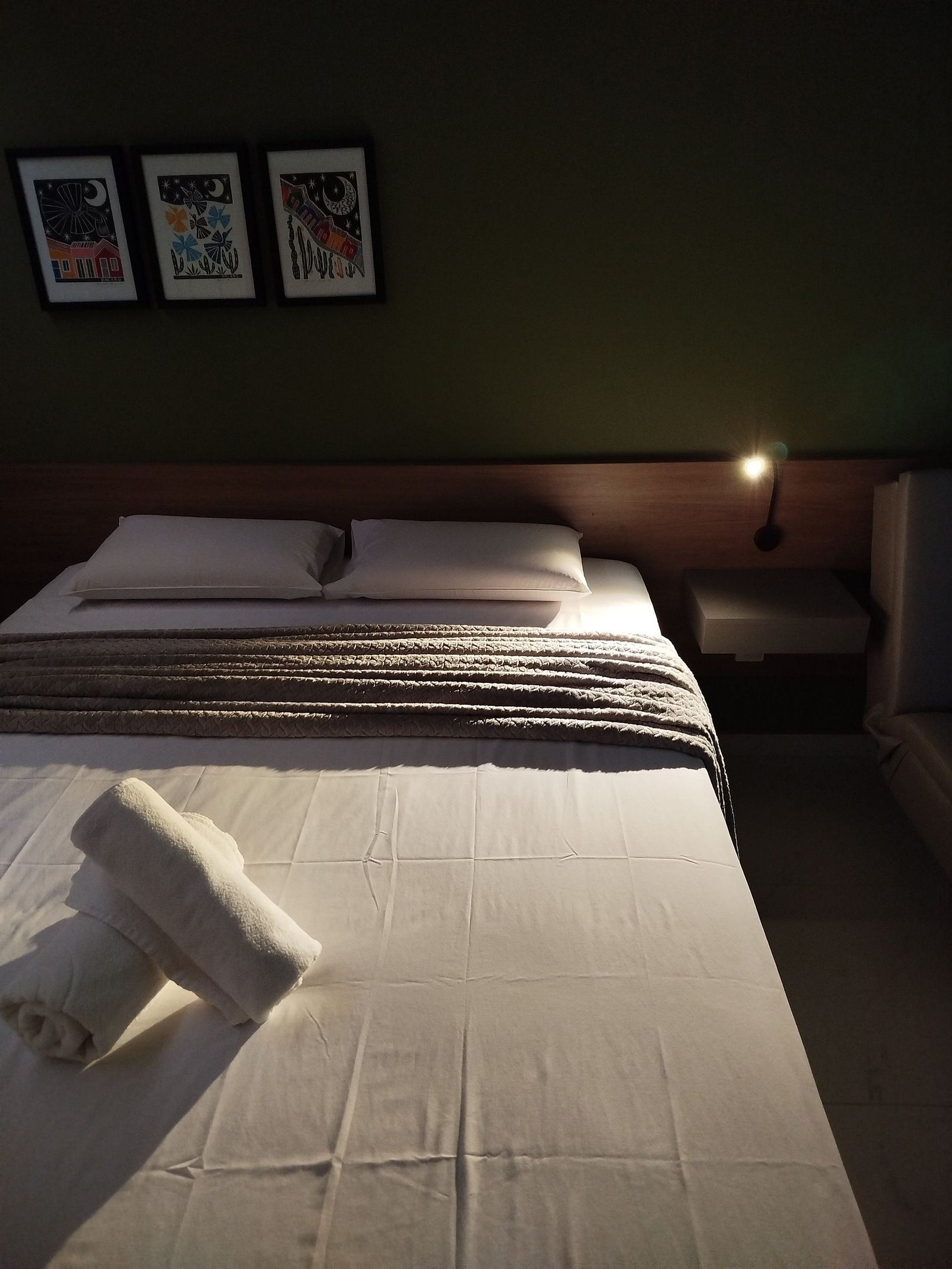 A neatly made bed with white linens and a rolled towel, a wooden headboard, a nightstand with a lamp, and three framed artworks on the wall.