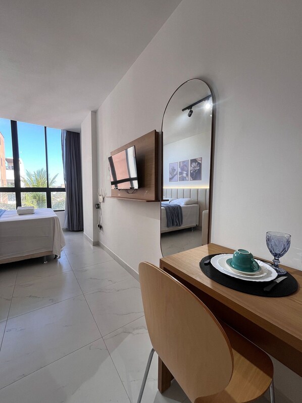 A hotel room with a bed, a desk with a chair, a dining table with a cup and glass, a large mirror, and a TV on the wall. - Porto City - Porto de Galinhas (Ipojuca)