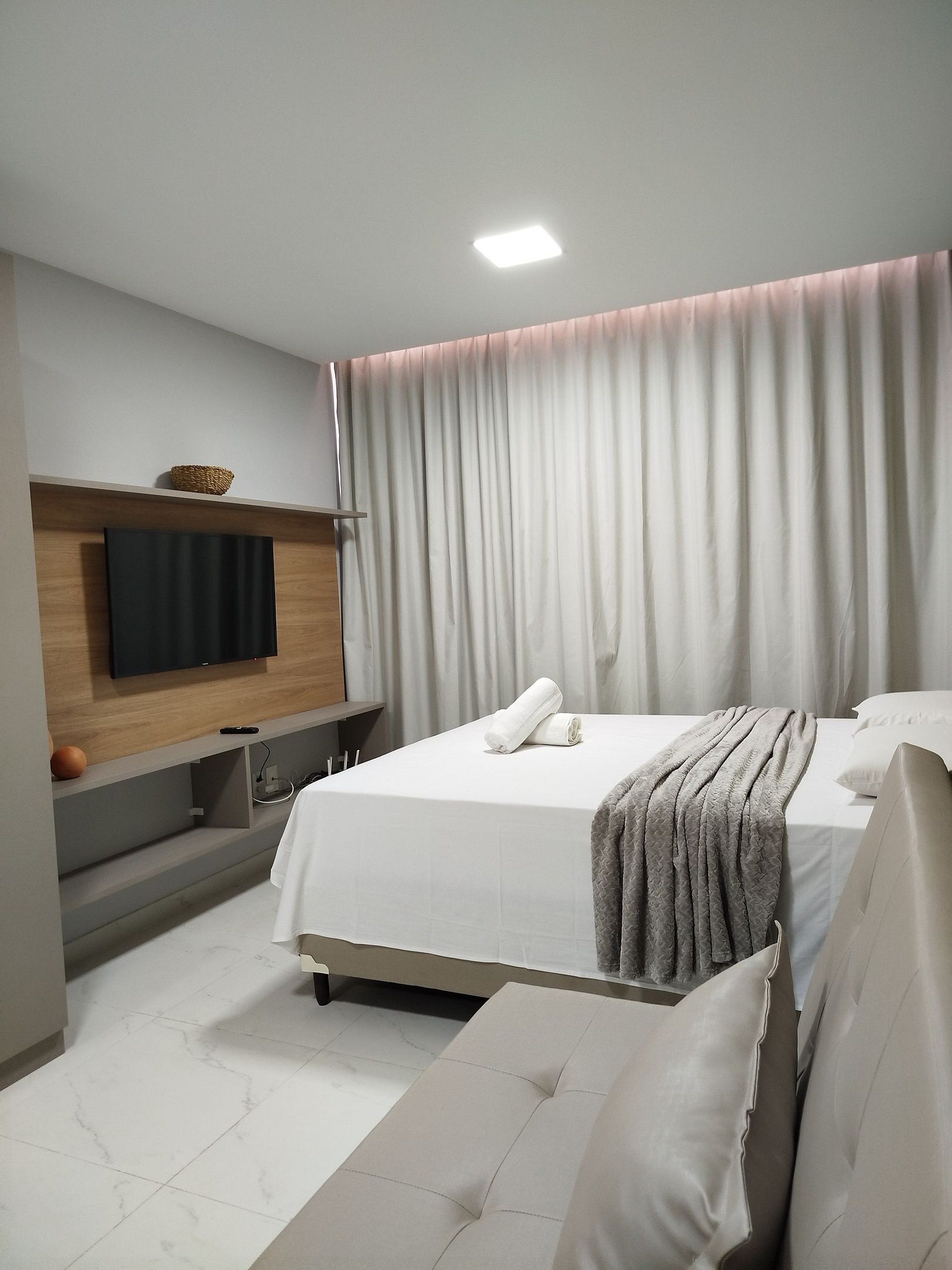 Quarto de hotel moderno com uma cama grande, uma televisão de tela plana na parede, uma mesinha de cabeceira e uma janela ampla com cortinas.