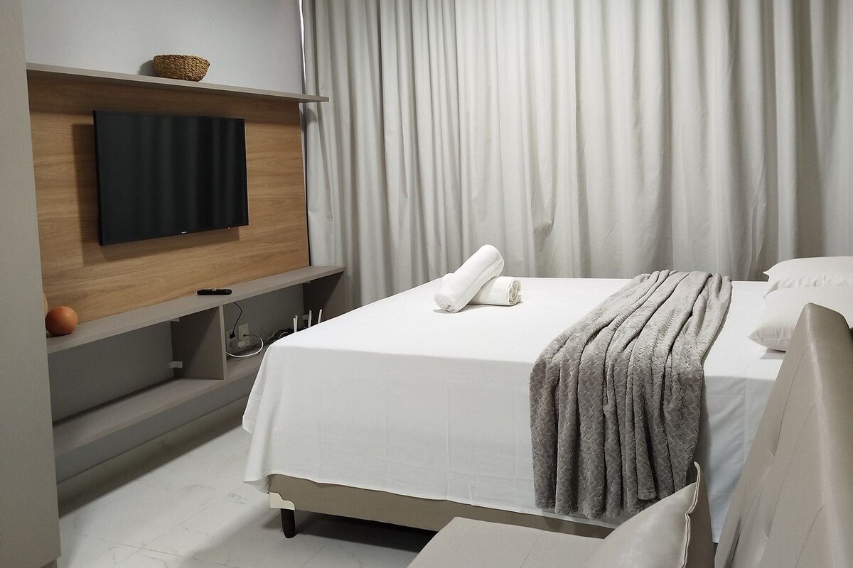 Habitación de hotel moderna con una cama grande, un televisor de pantalla plana montado en la pared, una mesita de noche y un ventanal con cortinas.