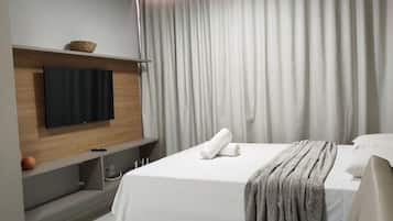 Quarto de hotel moderno com uma cama grande, uma televisão de tela plana na parede, uma mesinha de cabeceira e uma janela ampla com cortinas.