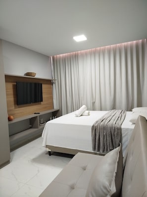Habitación de hotel moderna con una cama grande, un televisor de pantalla plana montado en la pared, una mesita de noche y un ventanal con cortinas.