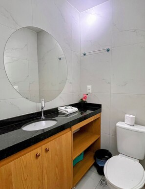 A bathroom with a round mirror, a sink, a toilet, and a shelf with a towel and a flower. - Porto City - Porto de Galinhas (Ipojuca)