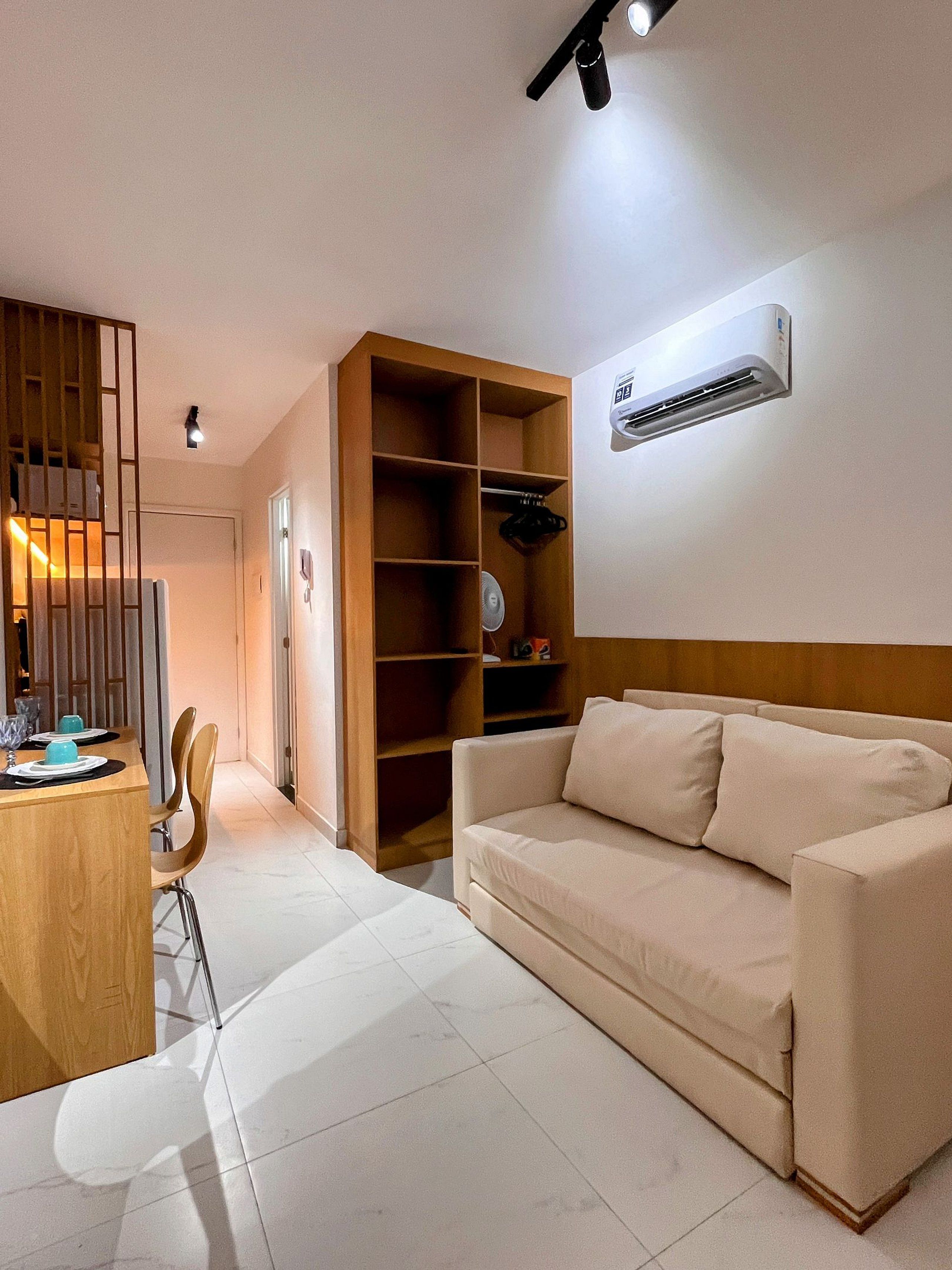 A living room with a beige sofa, a wooden dining table with chairs, a built-in shelving unit, and an air conditioning unit on the wall.
