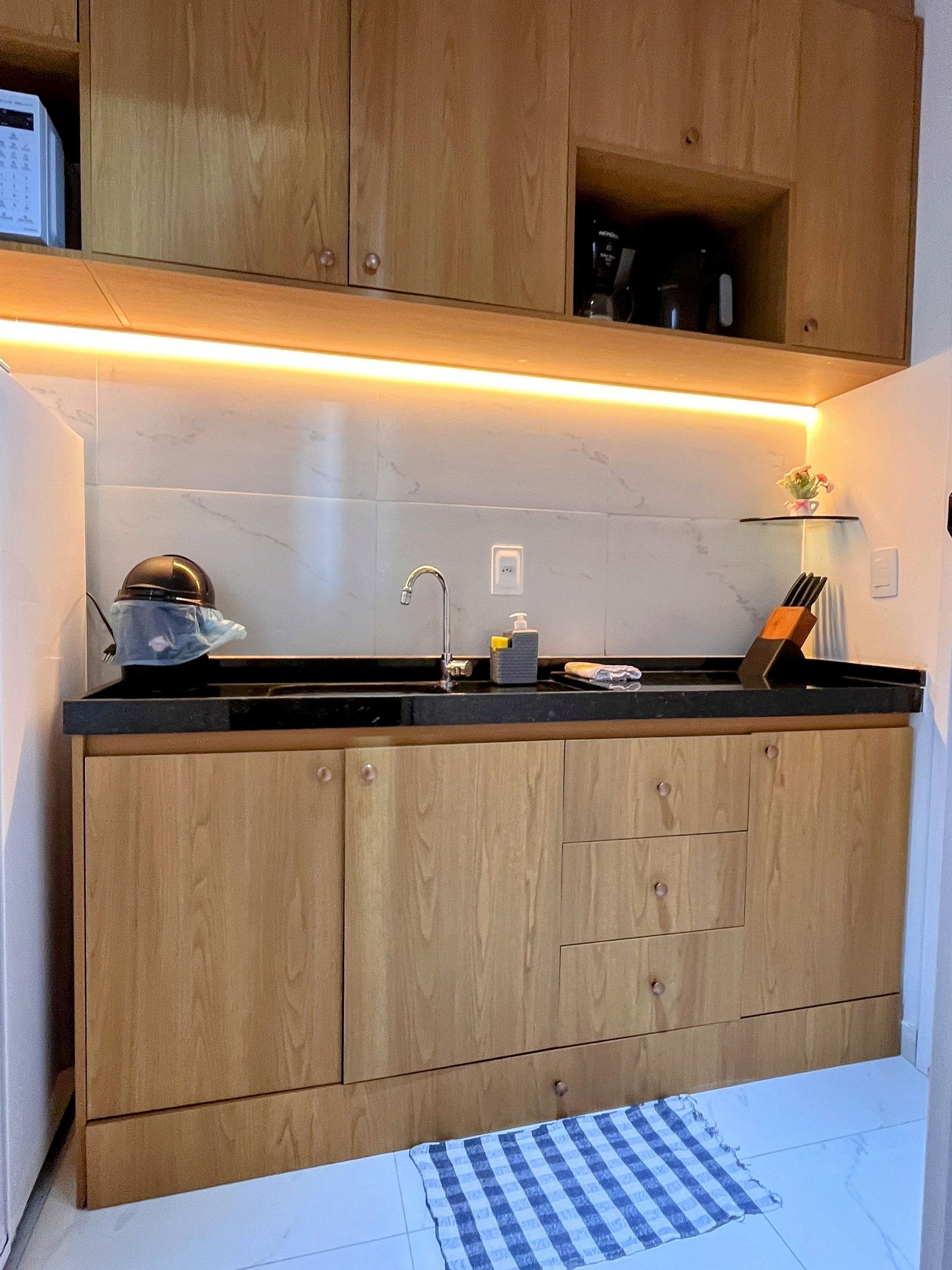 Une cuisine avec des meubles de rangement en bois, un plan de travail noir, un évier et une petite étagère sur laquelle sont posés divers objets.