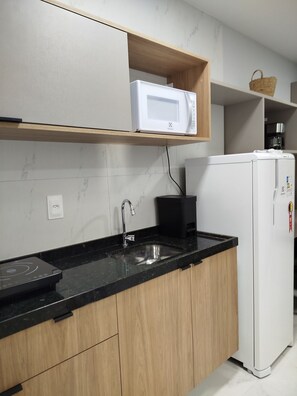 A kitchen with a black countertop, a white microwave, a white refrigerator, and a wooden shelf above the counter. - Porto City - Porto de Galinhas (Ipojuca)