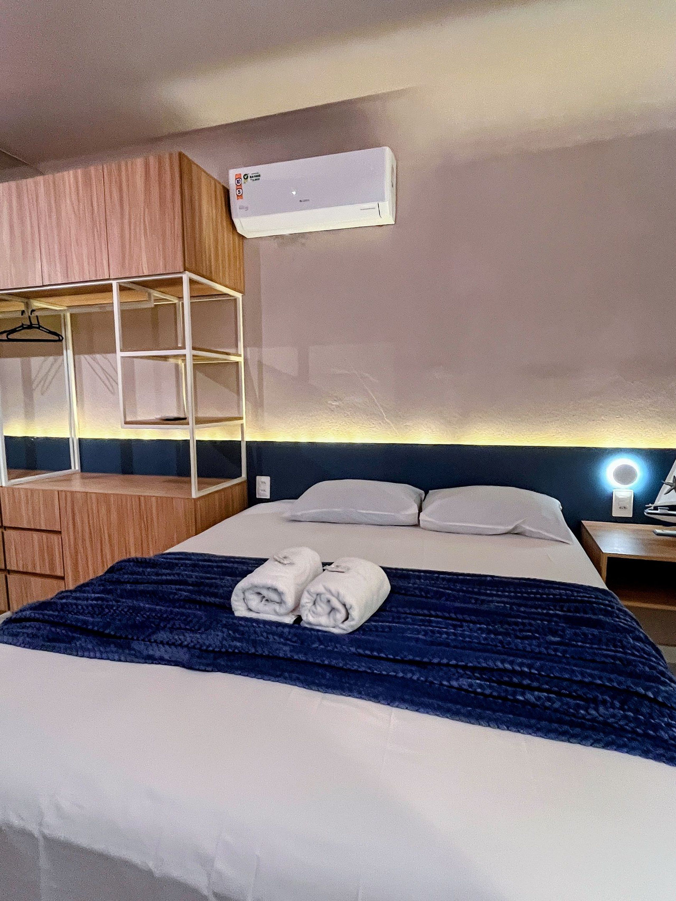 A hotel room with a bed, a hanging wardrobe, a bedside table with a lamp, and an air conditioning unit.