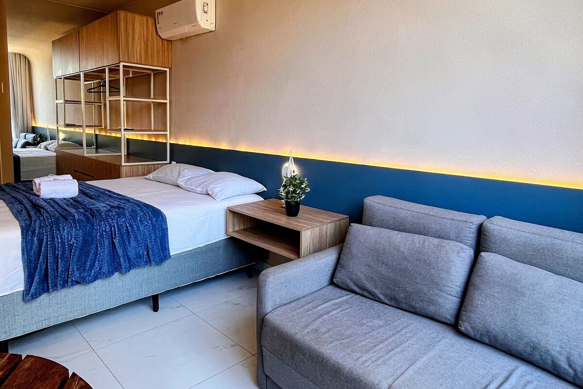 A hotel room with a bed, a sofa, a small table, and a wall-mounted air conditioner.
