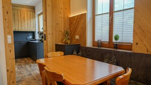 Une table de salle à manger en bois, avec des chaises, un coin cuisine et une fenêtre avec des stores.