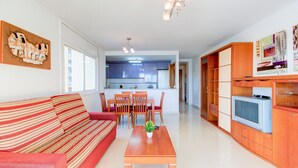 Una sala moderna con un sofá, una mesa de comedor con sillas, un televisor en un mueble de madera y una cocina al fondo.