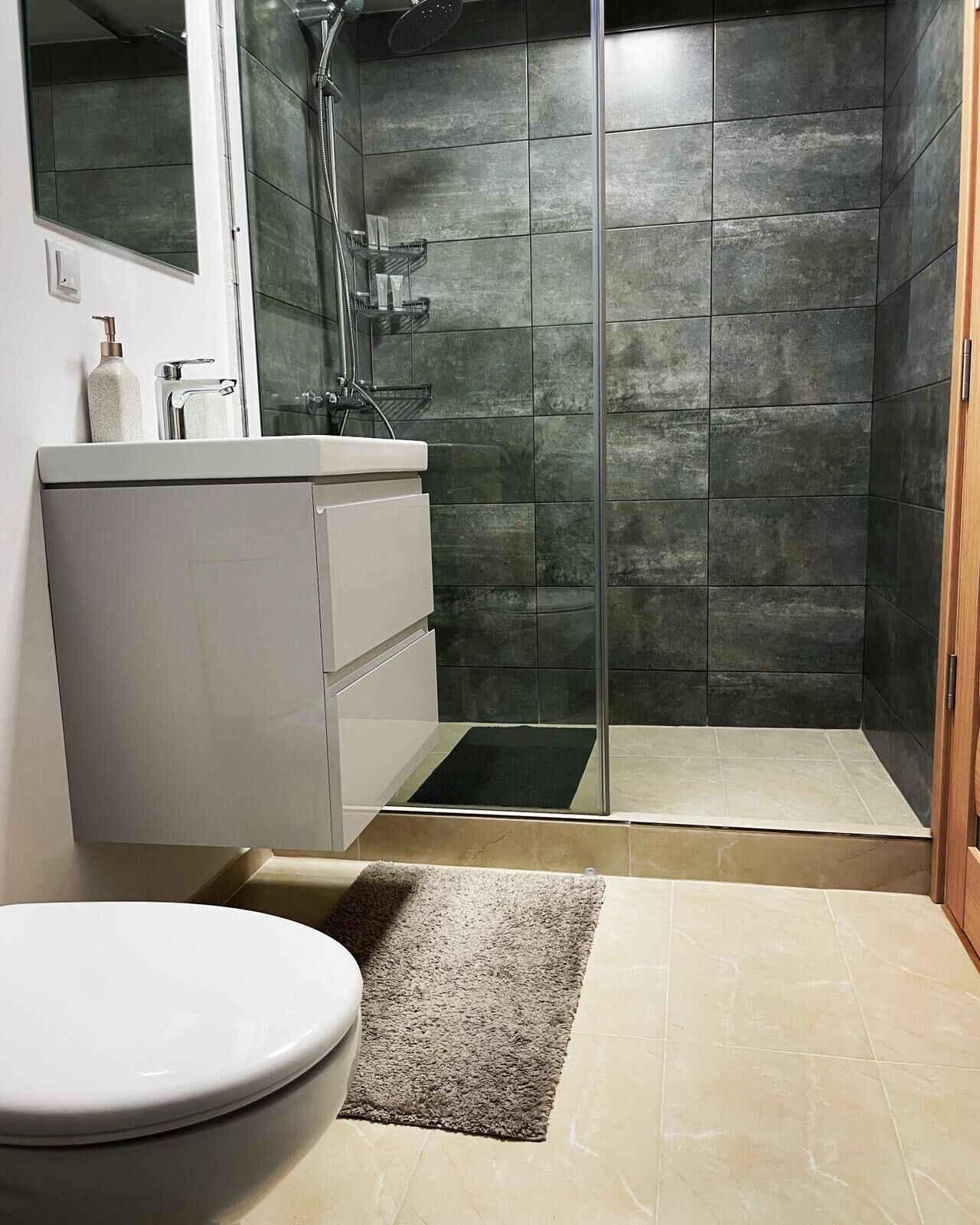 Un baño moderno con cabina de ducha de vidrio, un tocador blanco con espejo y piso de baldosas color beige.