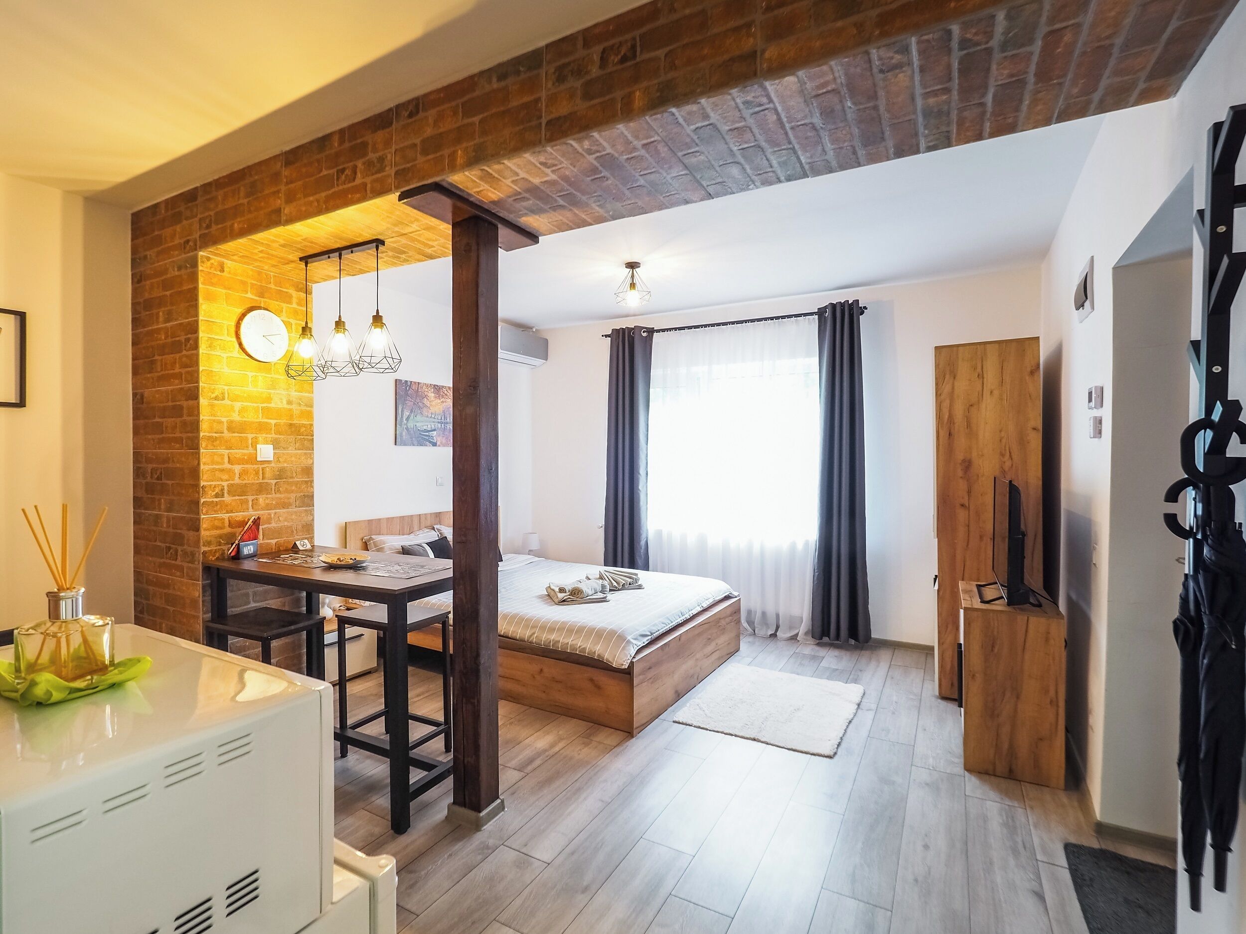 A modern bedroom with a brick wall, a wooden bed, a desk, and a dining area.