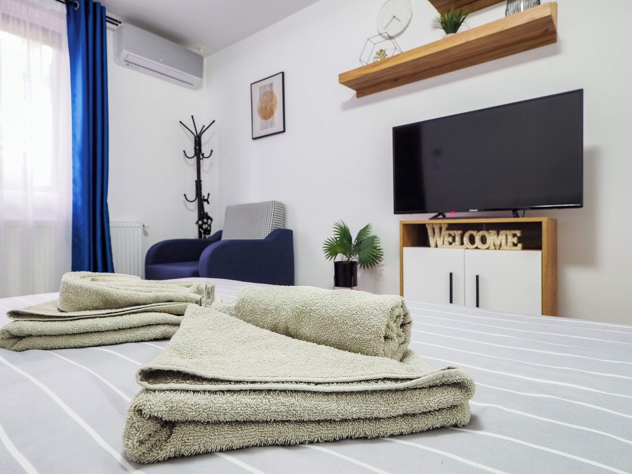 Una cama bien hecha con toallas dobladas, un televisor sobre un soporte de madera, una silla azul y un cartel que dice 'WELCOME'.