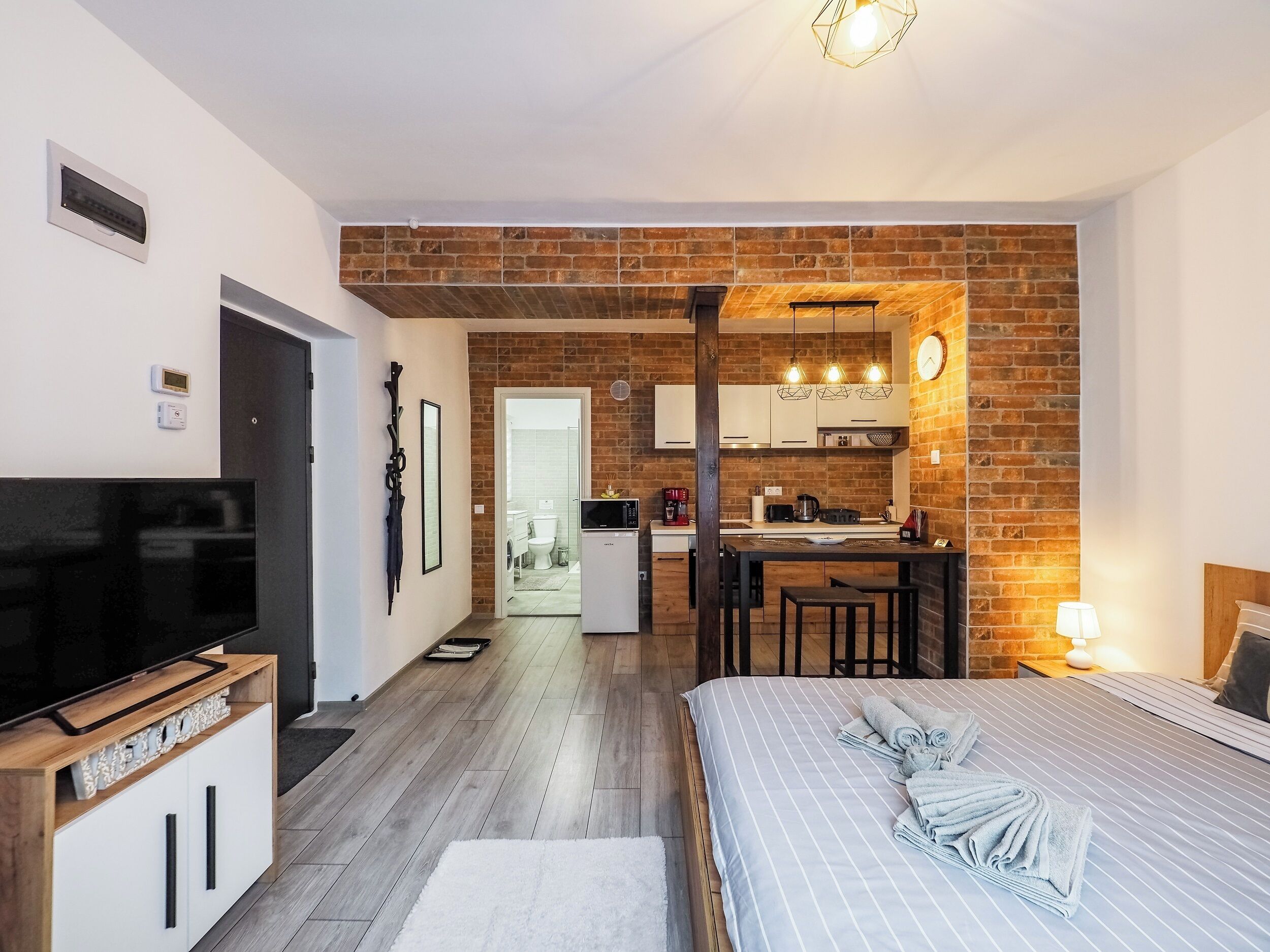 Ein modernes Hotelzimmer mit Fernseher, einem Holzschrank, einem Bett mit Handtüchern, einer Küchenzeile mit Backsteinwand und einem durch eine offene Tür einsehbaren Badezimmer.