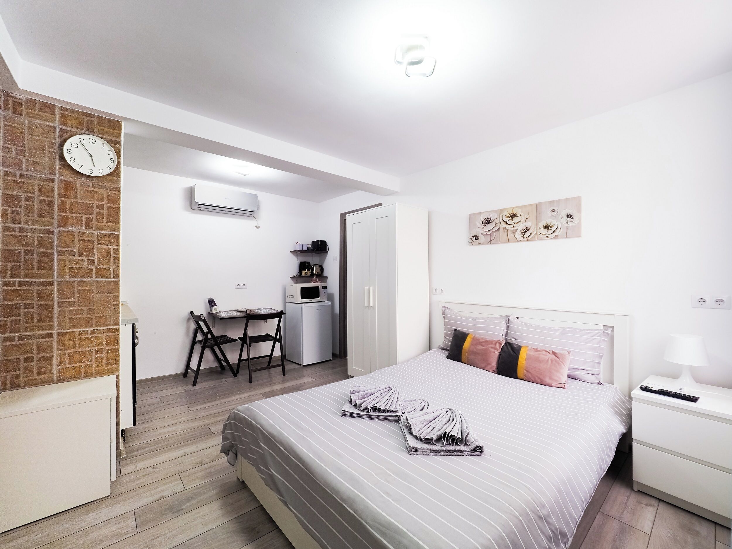 Un dormitorio con cama, mesita de noche, escritorio con silla, microondas, refrigerador y un reloj en la pared.