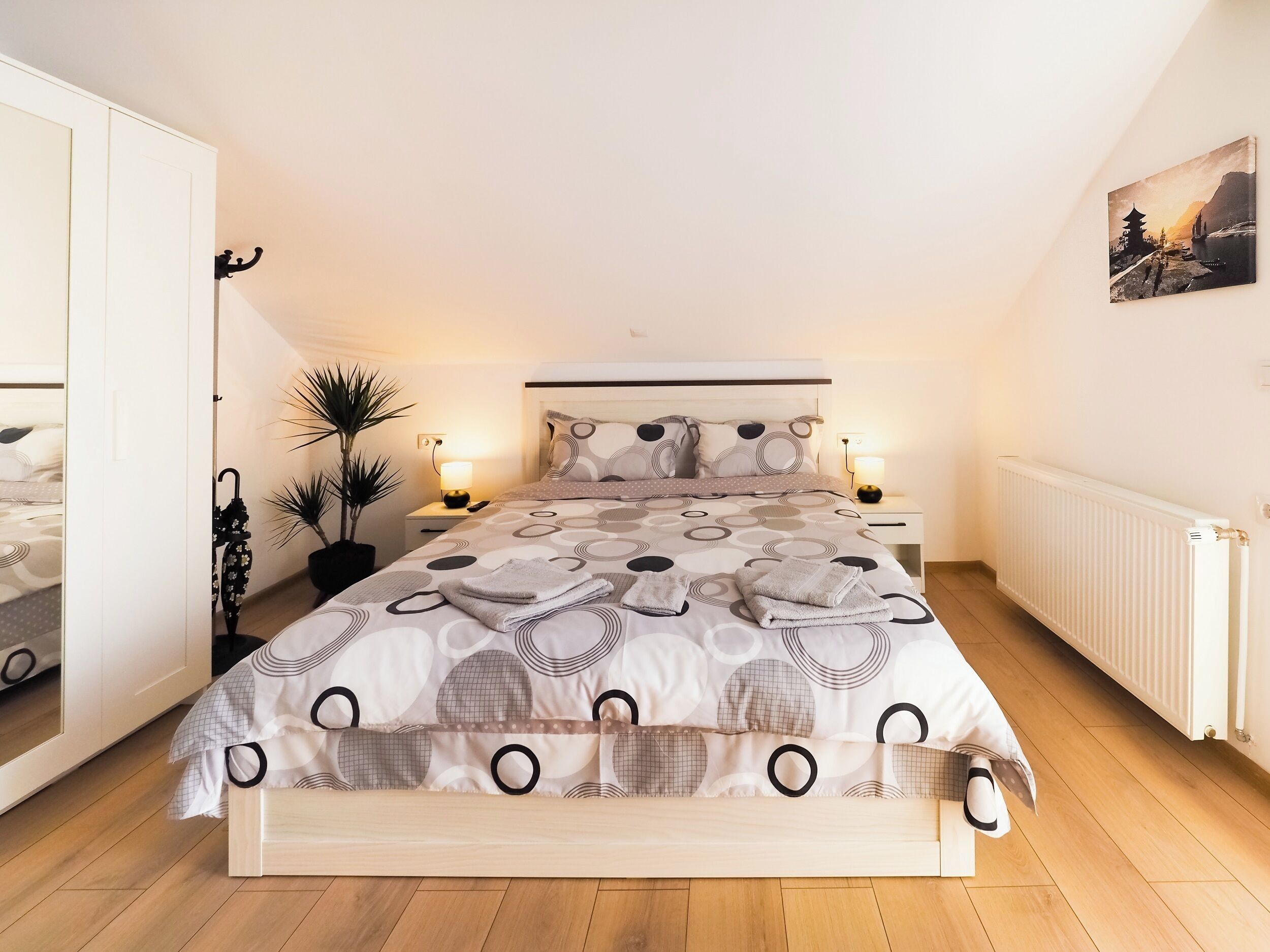 A neatly arranged bedroom with a bed, bedside tables, a radiator, and a wall-mounted painting.