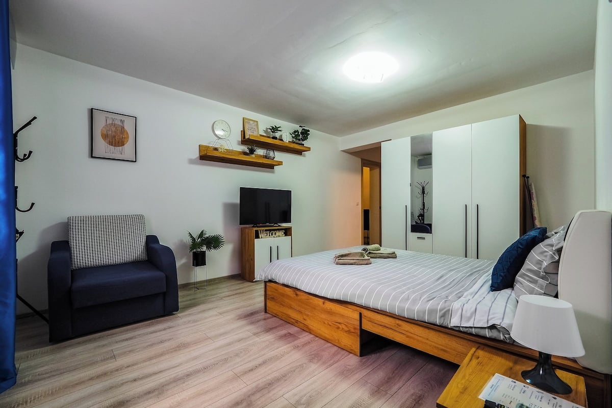 A bedroom with a bed, a TV, a wardrobe, a sofa, and a small table with a lamp.