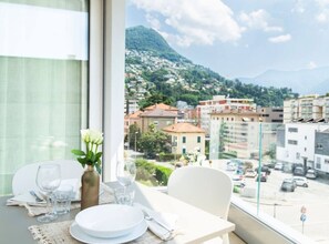 A dining area with a table set for two, a vase with flowers, and a view of a cityscape with buildings and a mountain in the background. - Elegant new Apt with AC, private balcony & garage (Viganello)