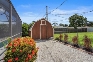 A small wooden shed with a door, surrounded by gravel and a flower bed with red flowers.