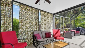Un patio iluminado por el sol con cerramiento de vidrio, mobiliario rojo y gris, y una pared con estampado de hojas tropicales.