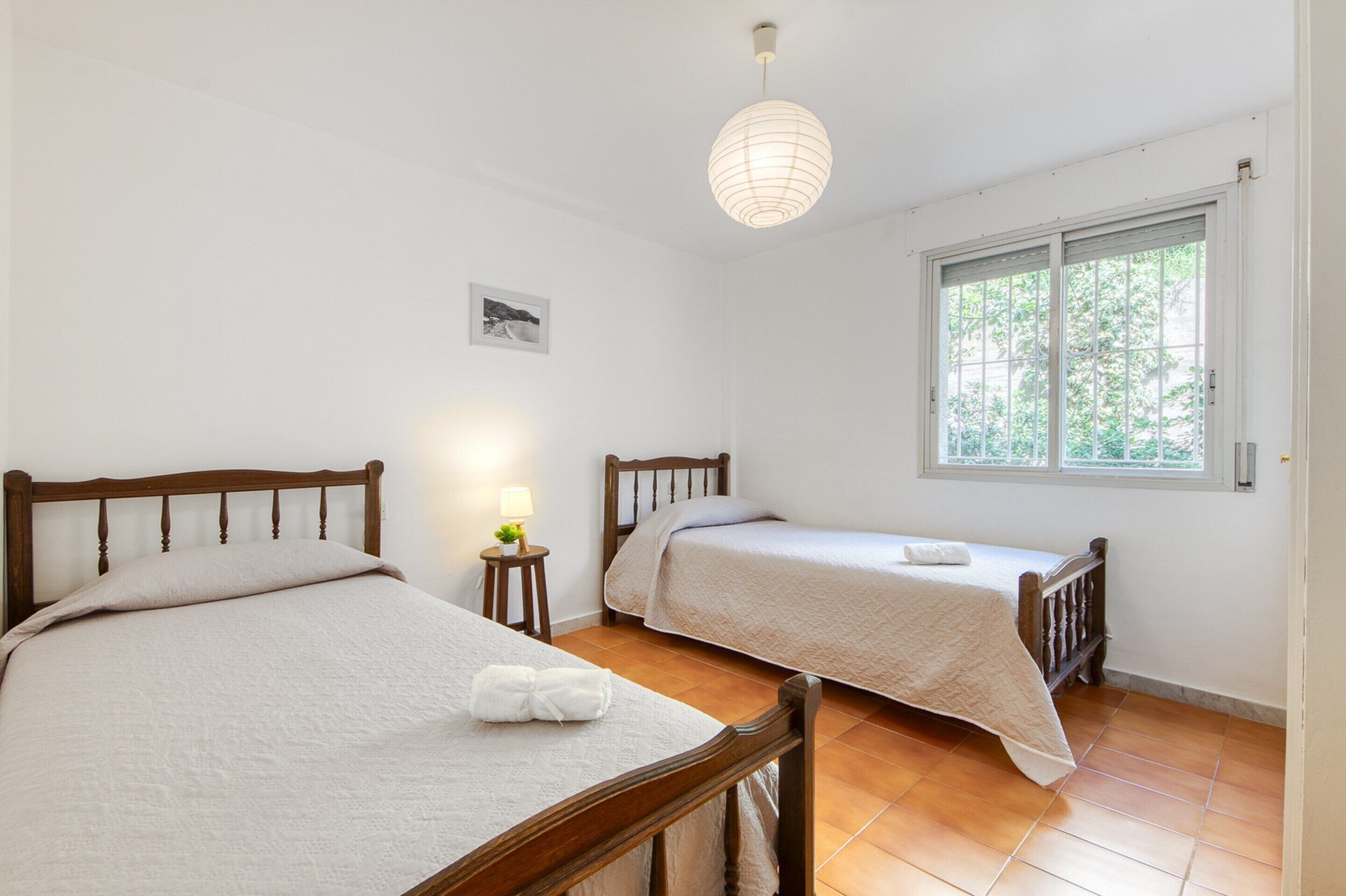 A room with two wooden beds, a small bedside table with a lamp, a hanging light fixture, and a window with a view of greenery.