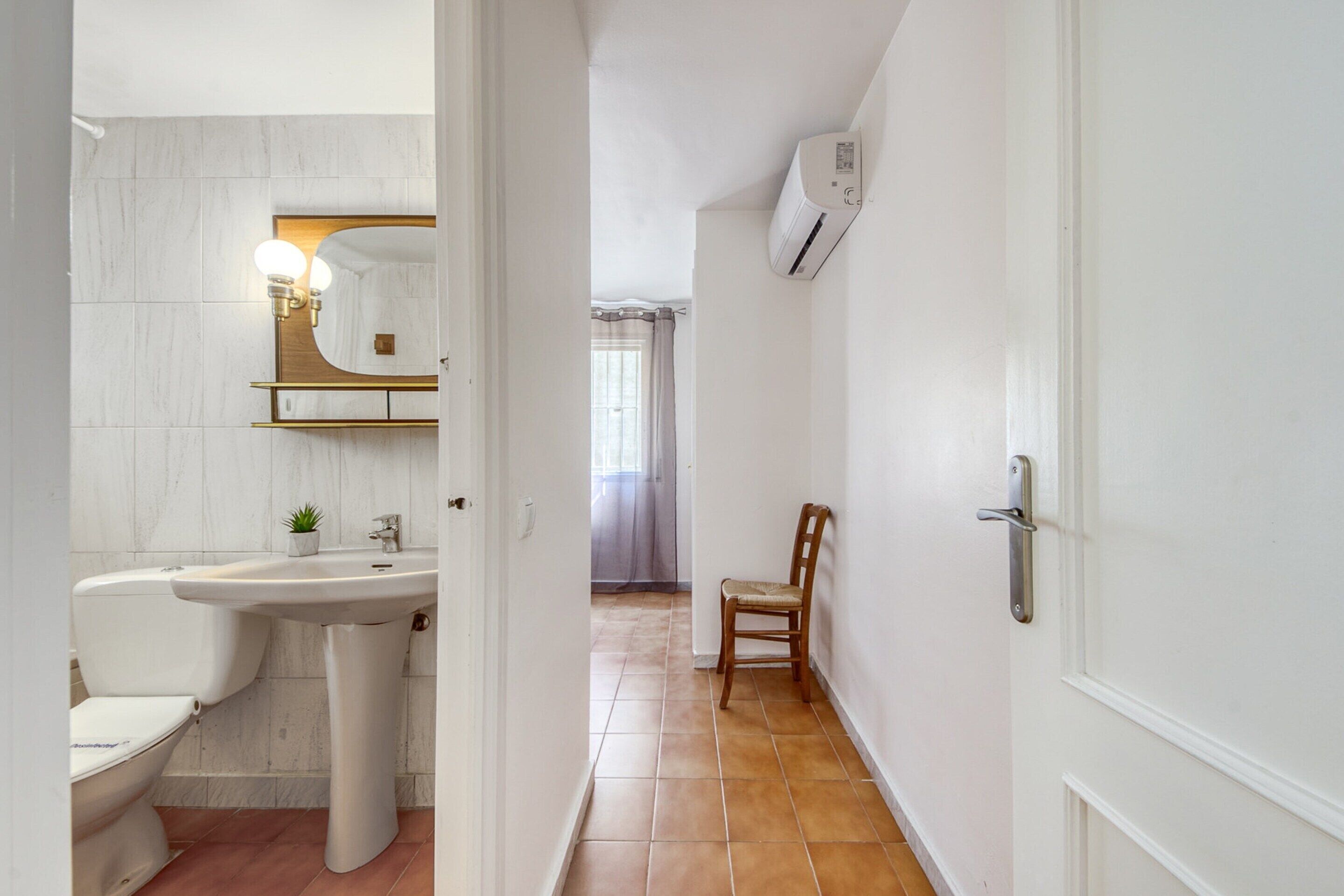 Une salle de bain avec un lavabo, des toilettes et un miroir blancs, une chaise en bois et un appareil de climatisation fixé au mur.