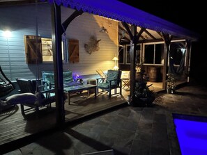 A covered outdoor seating area with chairs and tables, a wooden wall, and a small pool.