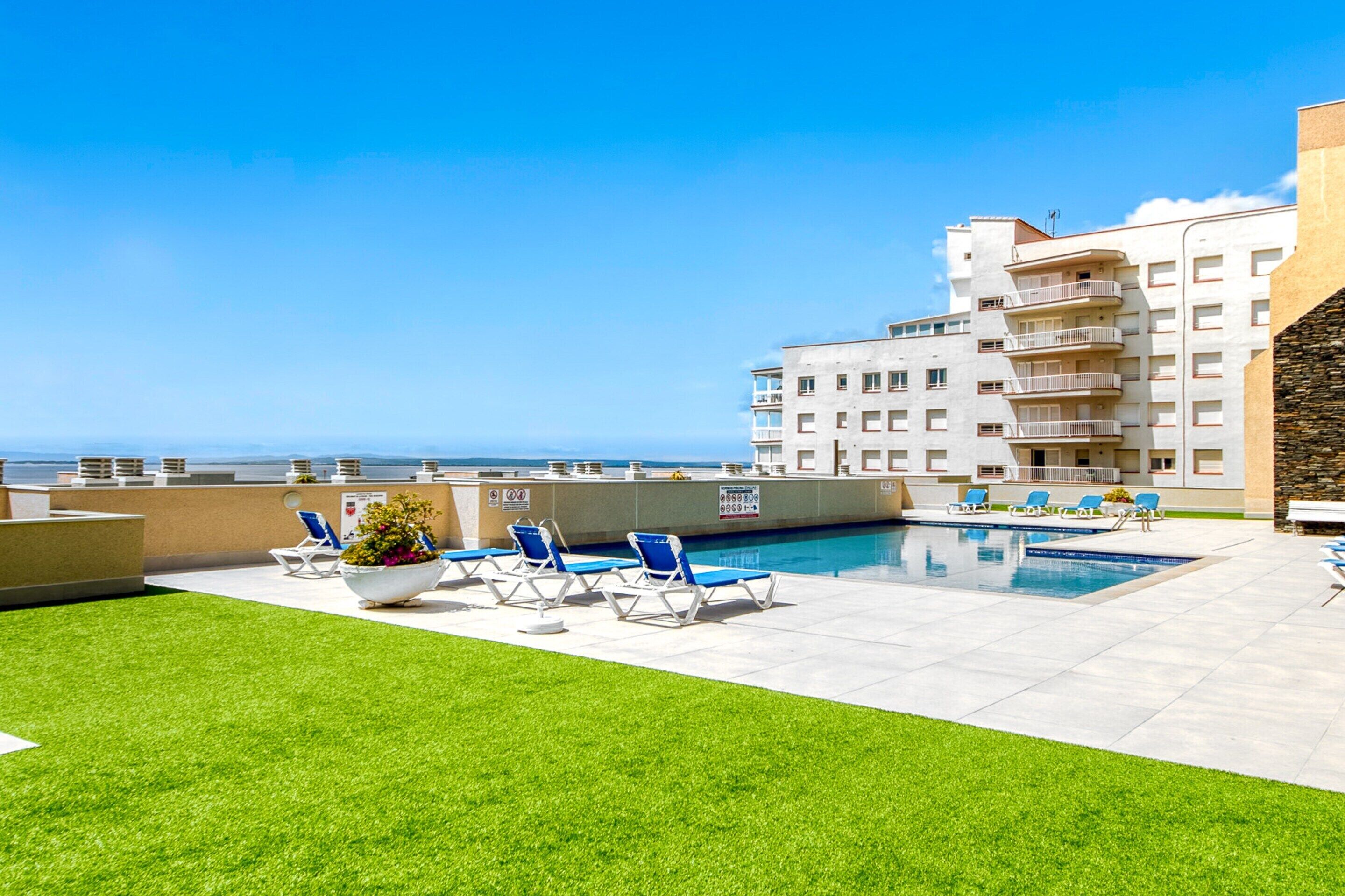 A rooftop pool area with a clear view of the ocean, surrounded by a building with multiple balconies and a well-maintained lawn.