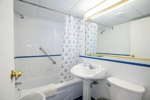 A bathroom with a bathtub, a sink, and a toilet. There is a shower curtain with anchor patterns, a mirror above the sink, and a blue stripe on the wall. - Dorchester Cosy Studio (Montreal)