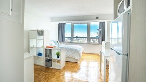 Une chambre moderne avec un lit, un bureau, une chaise, une petite plante et une vue sur la ville grâce à de grandes fenêtres.