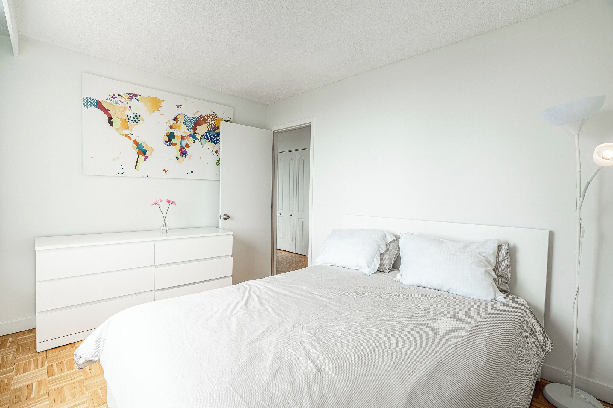A modern bedroom with a large bed, a dresser, a world map artwork, and a floor lamp.