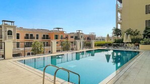 Una piscina con plataforma de baldosas, rodeada de edificios con balcones y un cielo despejado.