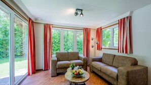 A spacious living room with a large window, a wooden floor, and a round coffee table with fruit. - Chalet Near De Hoge Veluwe National Park (Scherpenzeel)