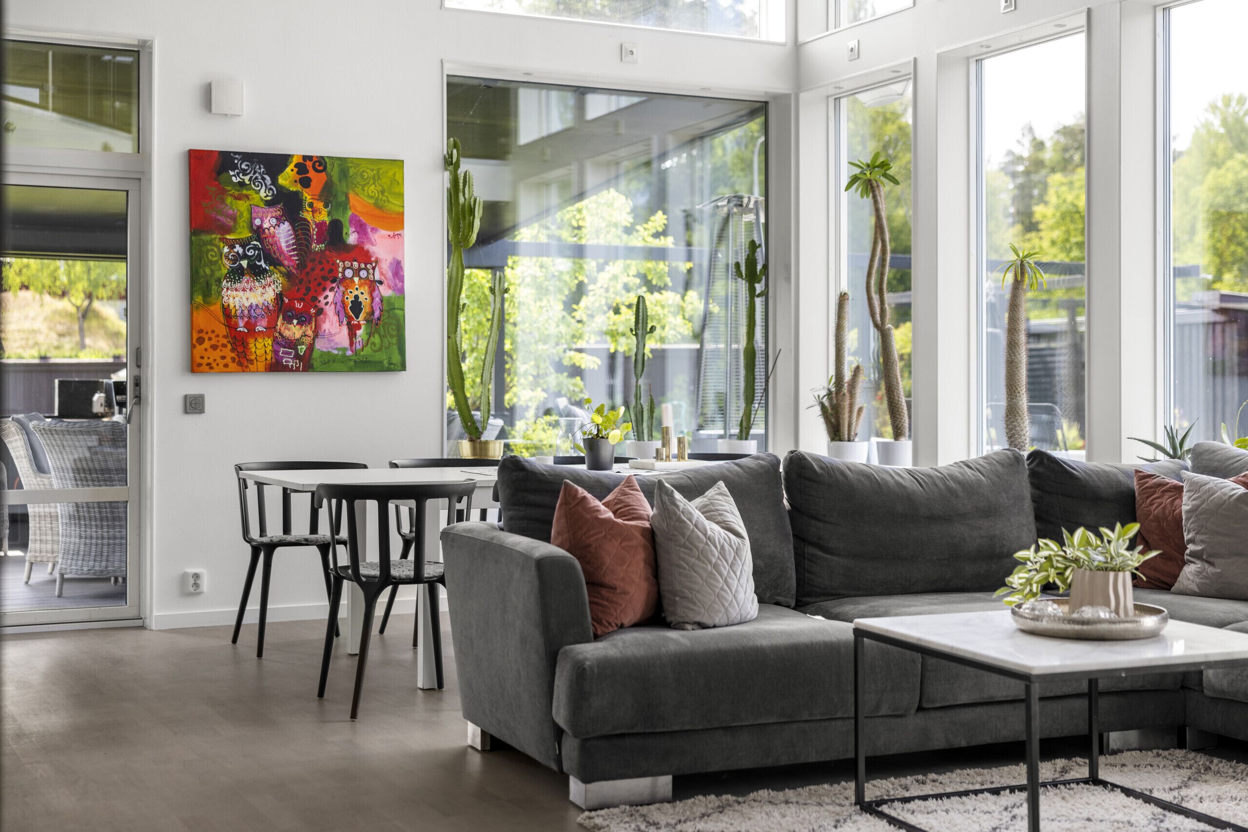 A modern living room with a grey sofa, a coffee table, and a dining area with a table and chairs. There is a large window with plants and a colorful painting on the wall.