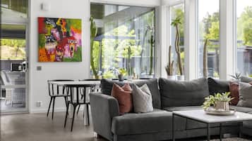 A modern living room with a grey sofa, a coffee table, and a dining area with a table and chairs. There is a large window with plants and a colorful painting on the wall.