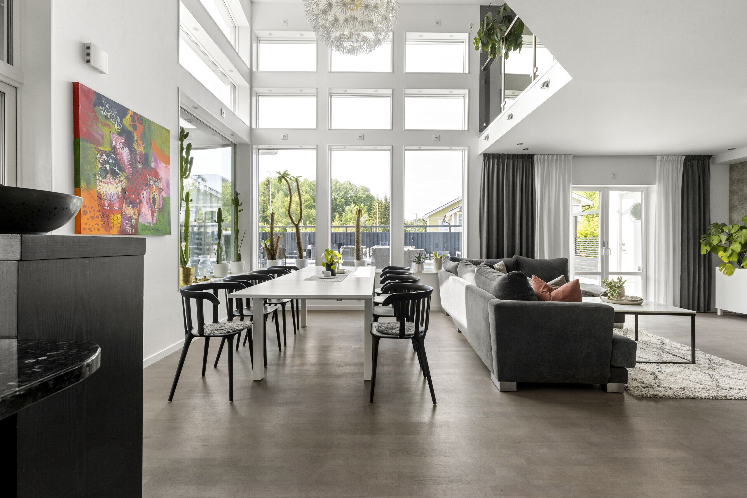 Una sala de estar moderna con un sofá gris, comedor con una mesa larga y sillas, y ventanas amplias que ofrecen vistas a la vegetación.