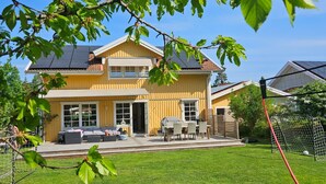 Ein zweistöckiges Haus mit großem Vorgarten, einer Terrasse mit Gartenmöbeln und einem sichtbaren Tennisplatz.
