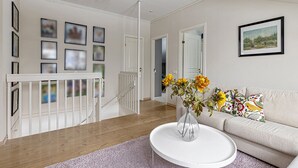 Ein geräumiges Wohnzimmer mit einem weißen Sofa, einem runden Couchtisch mit Blumen in einer Vase, einer Treppe mit weißer Geländer und gerahmten Bildern an den Wänden.
