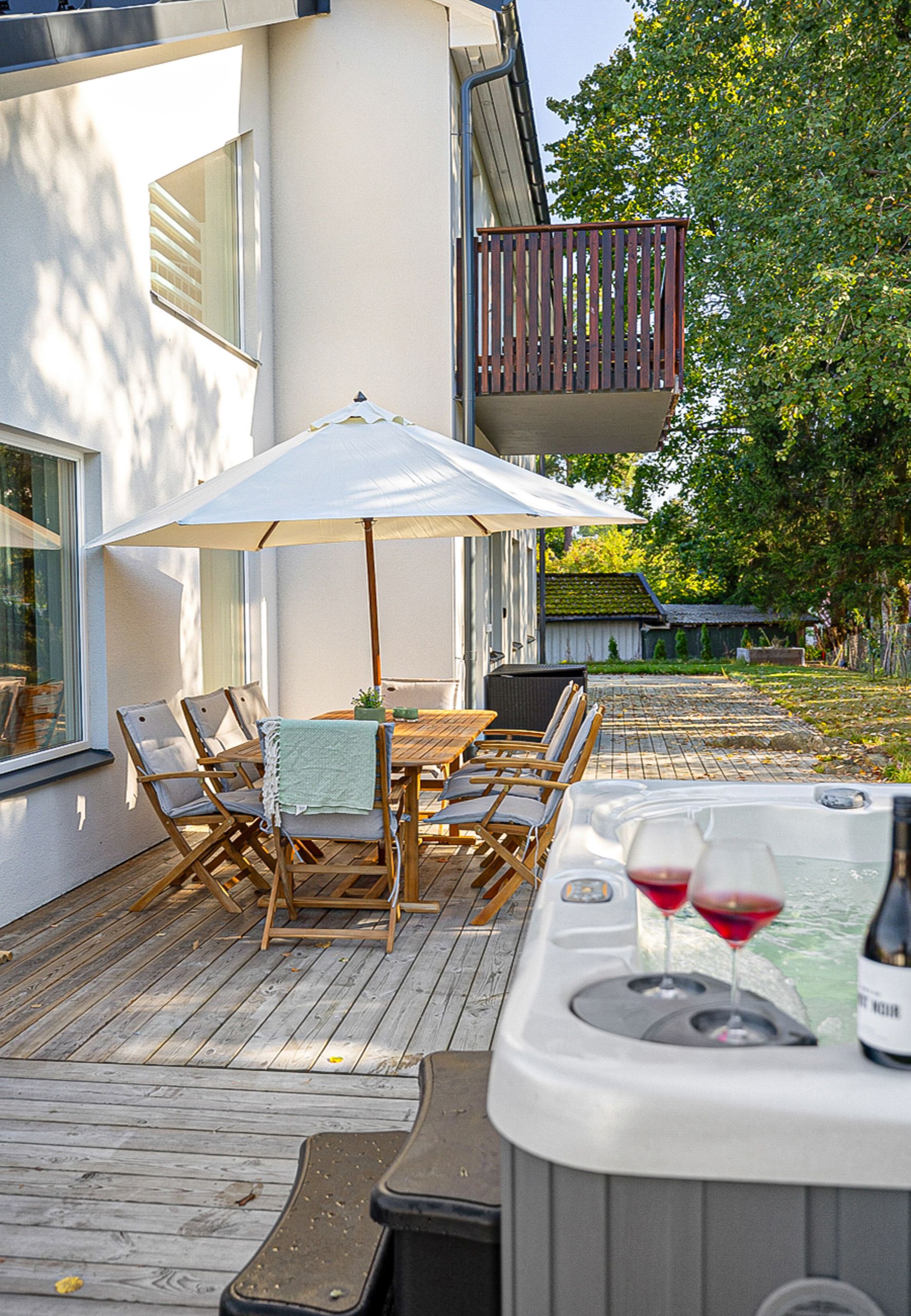 Un patio exterior con una mesa y sillas de madera, una sombrilla blanca y un jacuzzi con dos copas de vino tinto.