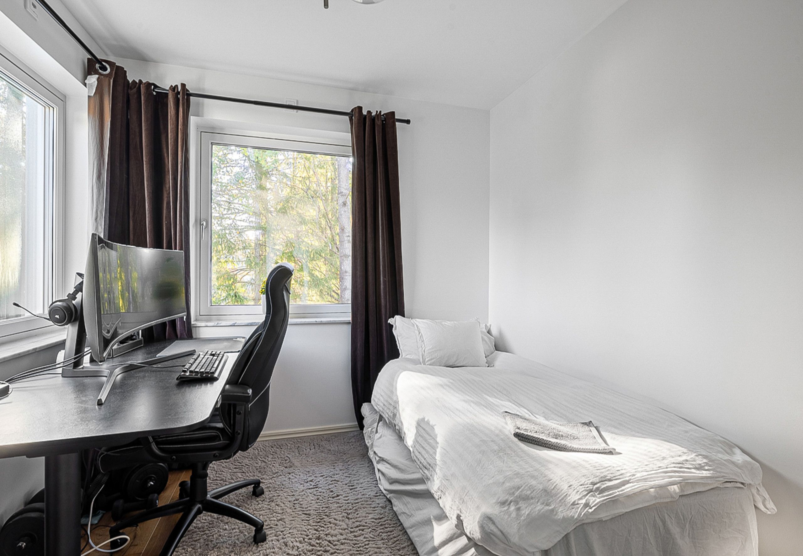 A bedroom with a single bed, a desk with a computer, and a window with curtains.