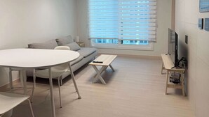 Ein modernes Wohnzimmer mit einer Couch, einem runden Tisch, einem Fernseher und einem Fenster mit Jalousien.
