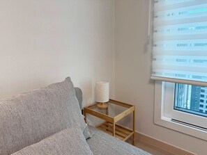 A room with a sofa, a small wooden side table with a lamp, and a window with blinds.