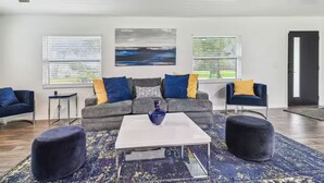 A modern living room with a grey sofa, blue and yellow cushions, a white coffee table, and a blue vase. There are two ottomans, a small side table, and a painting on the wall. - Modern 3bed, 2bath House in Wesley Chapel, with EV Charging Outlet, AC, & WiFi.  (Zephyrhills)