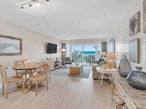 Un salon spacieux comprenant un ensemble de salle à manger en bois, un canapé et une table basse. Un téléviseur est fixé au mur, et des tableaux encadrés ornent les murs.